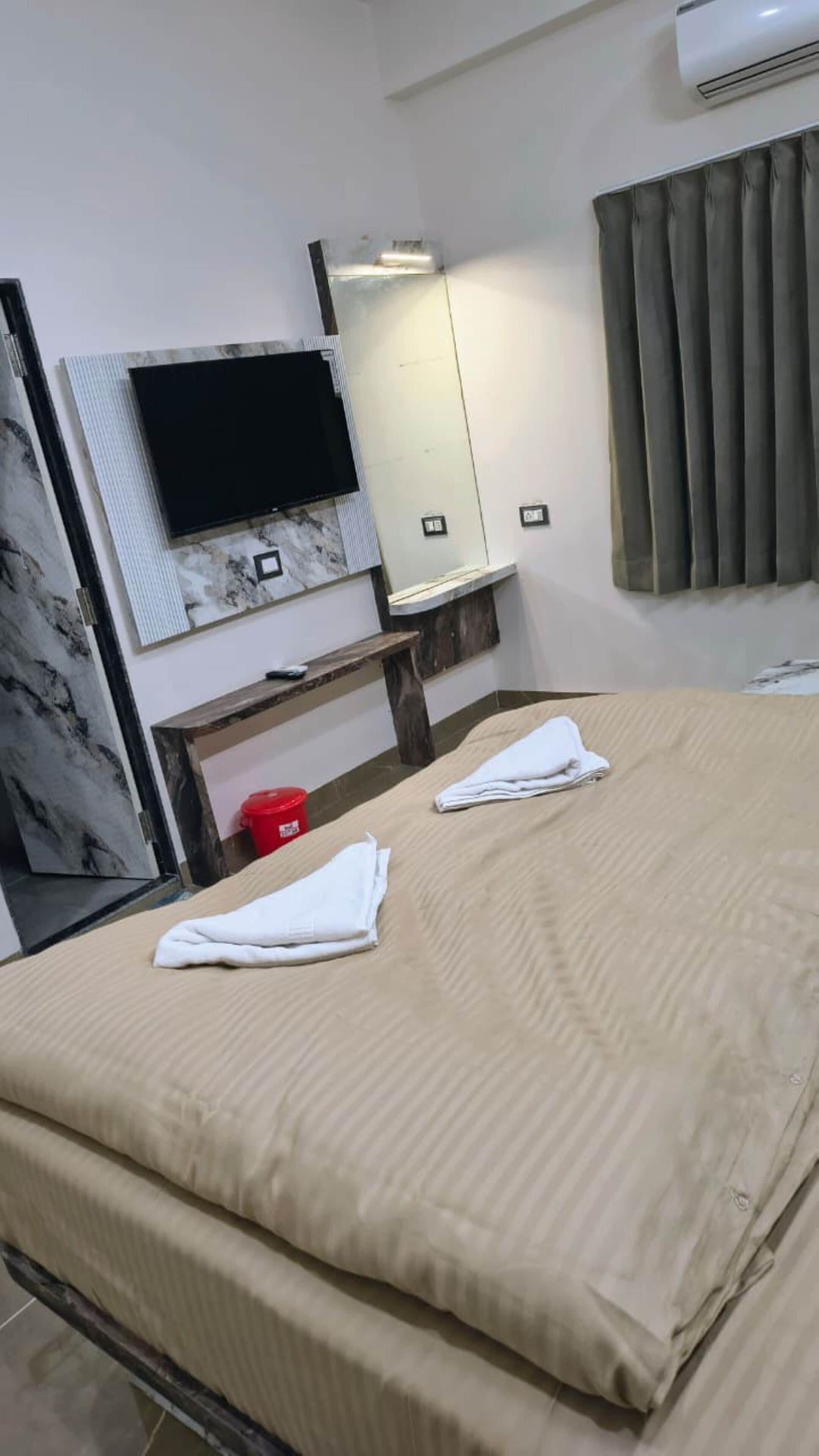 A hotel room with a bed, a flat-screen TV mounted on the wall, a mirror, and a window with curtains.