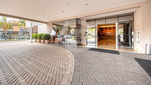 Entrada de un hotel con zona pavimentada en ladrillo, banco para sentarse, plantas en macetas y puerta de cristal que da al interior.
