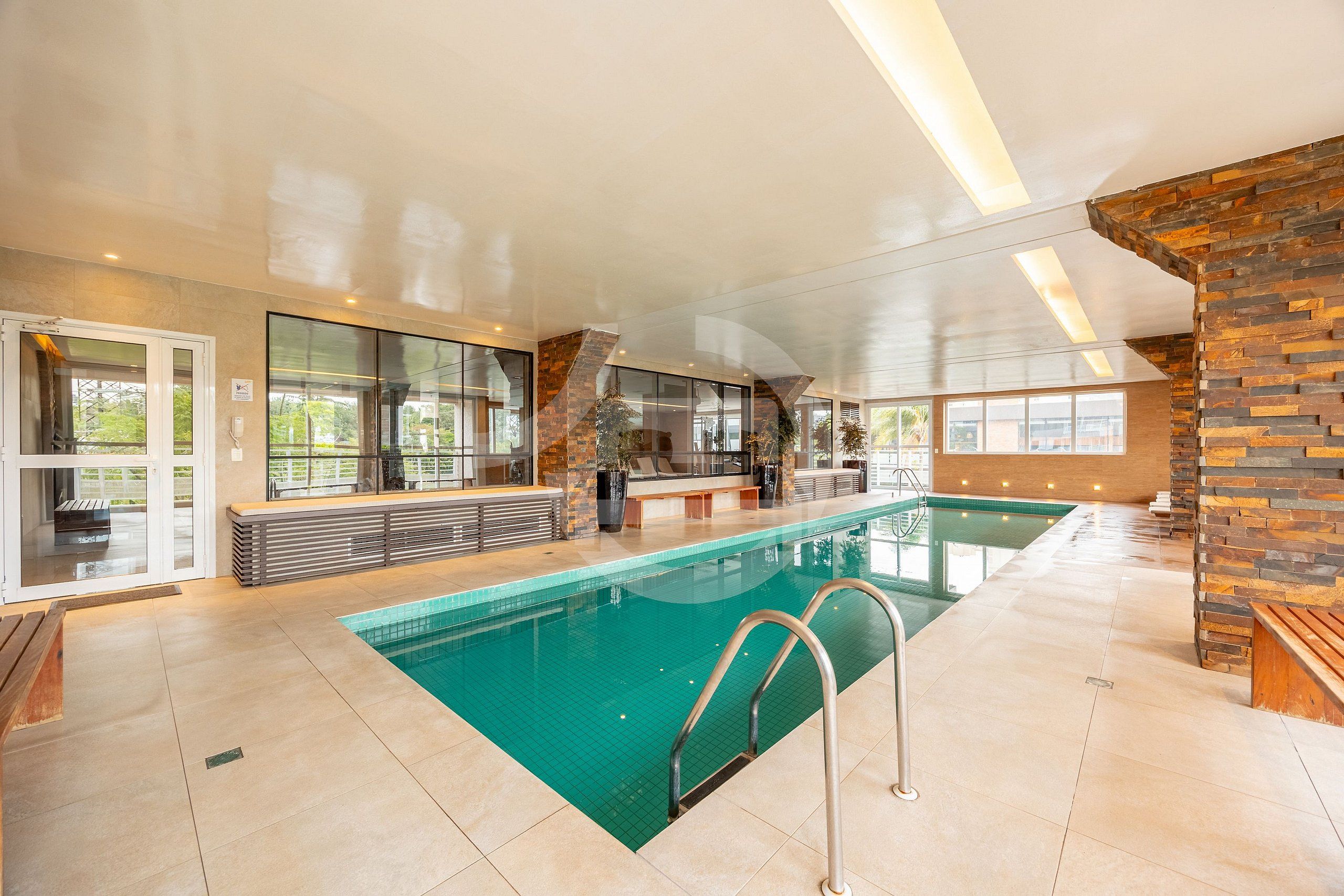 En indendørs swimmingpool med frit udsyn til bassinområdet, et loungeområde med en bænk, og en væg med detaljer i sten.