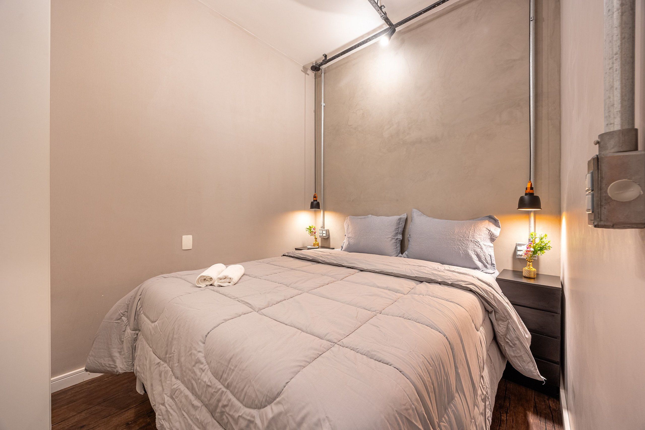 Una cama bien hecha con edredón gris, una mesita de noche con lámpara y un aplique en la pared.