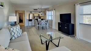 A modern living room with a sofa, a glass coffee table, and a TV. There is a kitchen in the background with white cabinets and a dining area with chairs. - Regency Towers #401 East | Gulf Front, Pool, Pickleball/Tennis (Pensacola Beach)