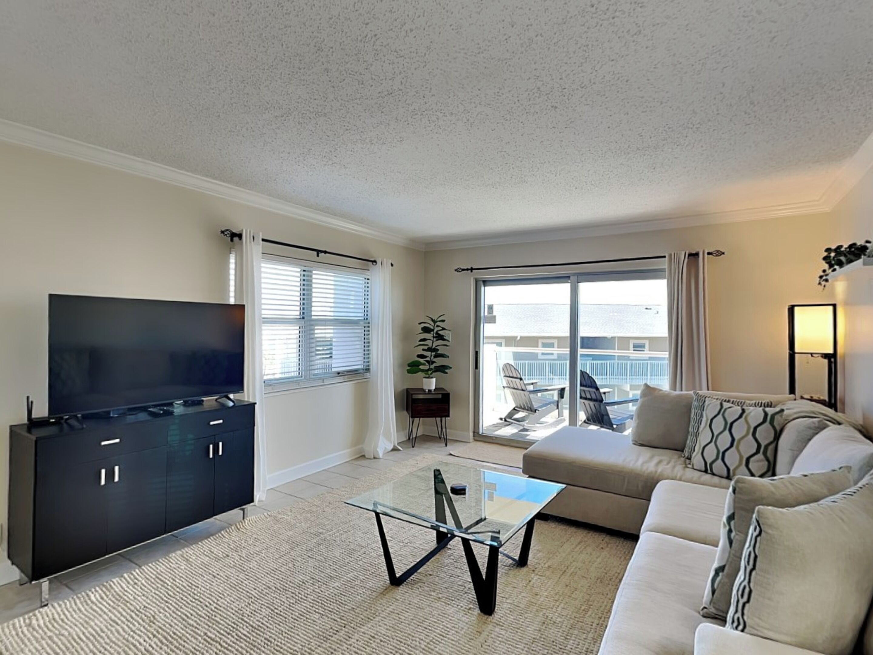 A living room with a flat-screen TV, a sofa, a glass coffee table, and a sliding glass door leading to a balcony with outdoor furniture.
