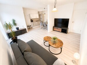 Una sala de estar moderna con un sofá, una mesa de centro y un televisor. Se ve una cocina al fondo.