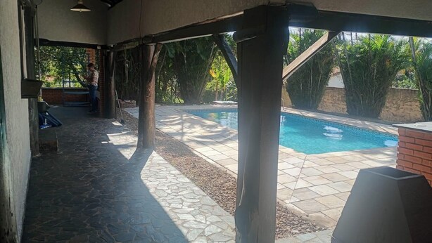 A covered outdoor area with a pool, stone flooring, and a person standing by a window. - Chácara Recanto da Coruja - São Pedro, SP (São Pedro)
