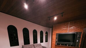 A spacious living area with a large flat-screen TV mounted on a brick wall, a grey sofa, and a brown couch. - Chácara Recanto da Coruja - São Pedro, SP (São Pedro)