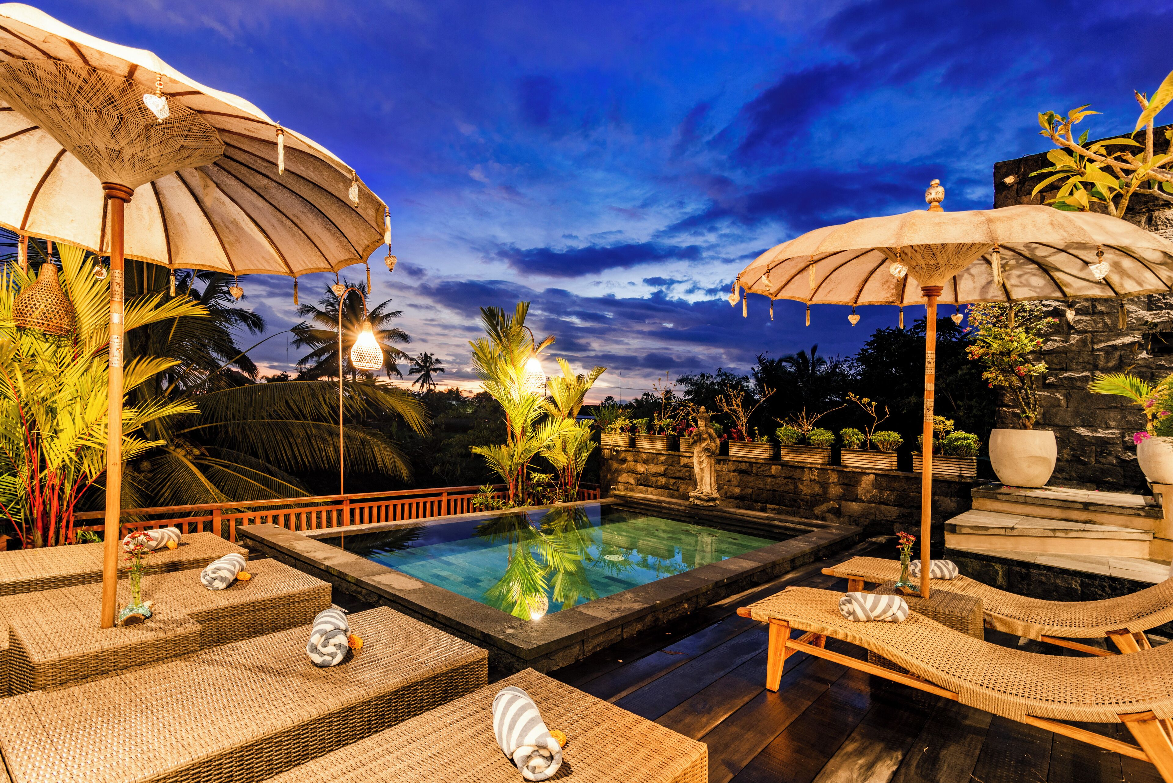 A pool area with lounge chairs and umbrellas, surrounded by tropical plants and a stone wall.
