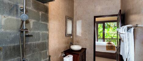 A bathroom with a stone shower, a sink, and a toilet. There is a window with a view of greenery outside.