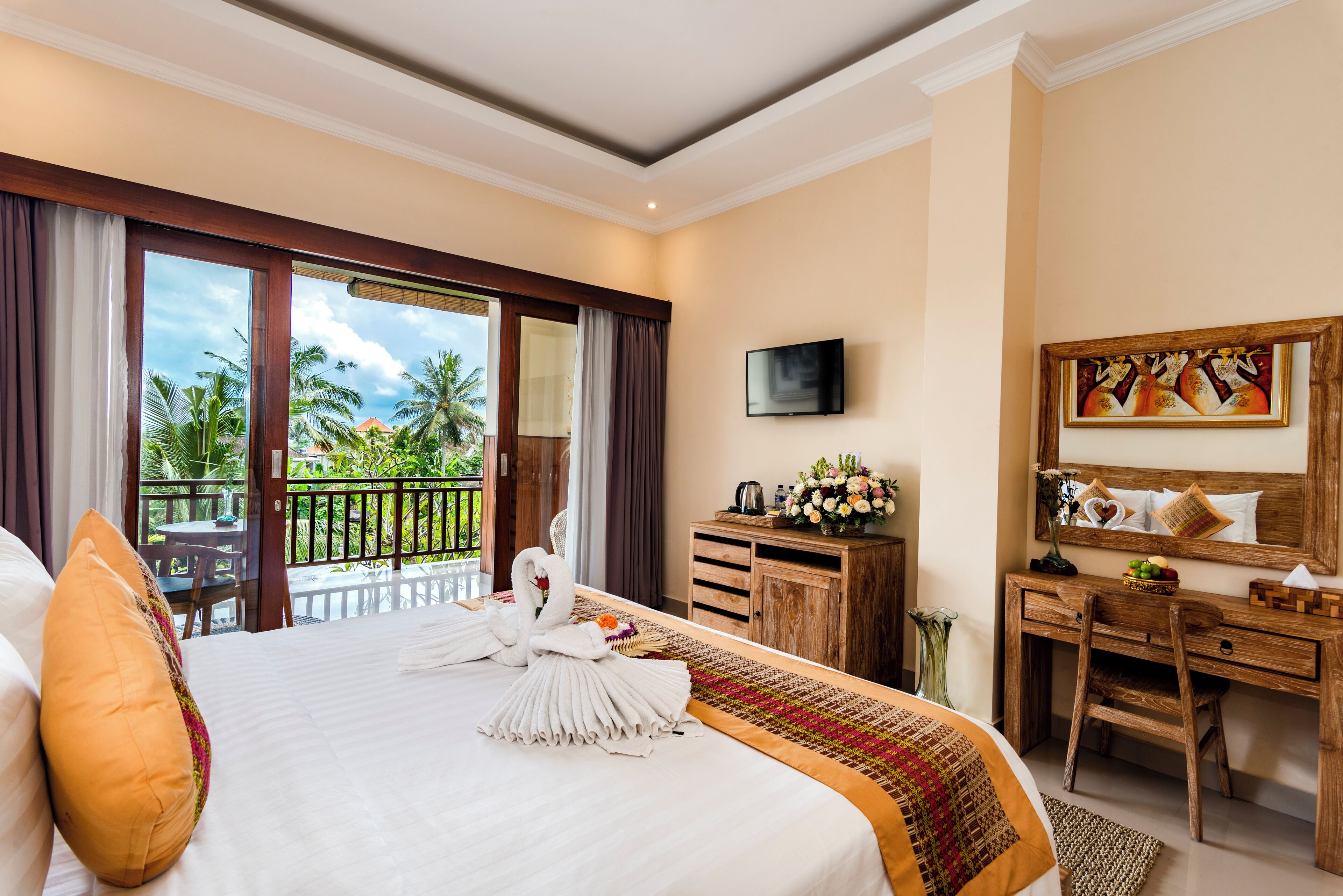 A hotel room with a large bed, a desk, a TV, and a balcony with a view of palm trees.