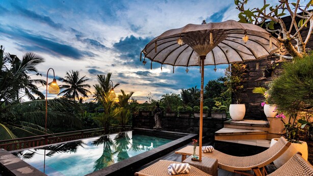 Un área junto a la piscina con sillas de descanso y una sombrilla, rodeada de plantas tropicales y una estructura de piedra.