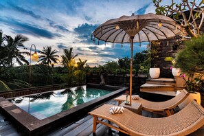 A poolside area with lounge chairs and an umbrella, surrounded by tropical plants and a stone structure. - Oka Suite Ubud by Dhananjaya Hospitality (Sebatu)