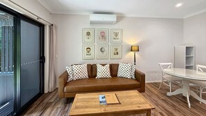 Een moderne woonkamer met een bruine leren bank, een houten salontafel en een glazen eettafel. Er hangen ingelijste foto’s aan de muur, staat een staande lamp en is er een witte boekenkast.