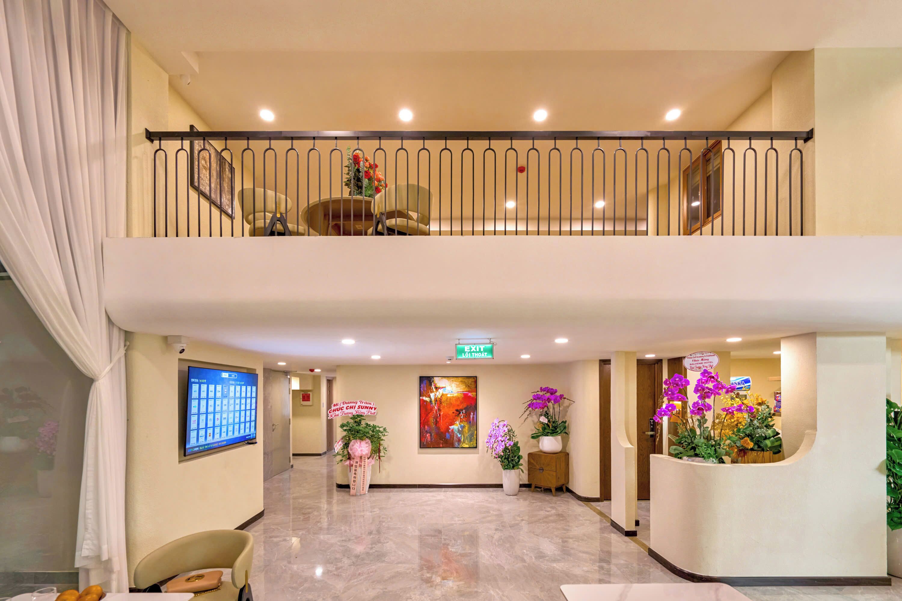 A hotel lobby with a reception desk, a TV screen, a staircase, and a large window with curtains.