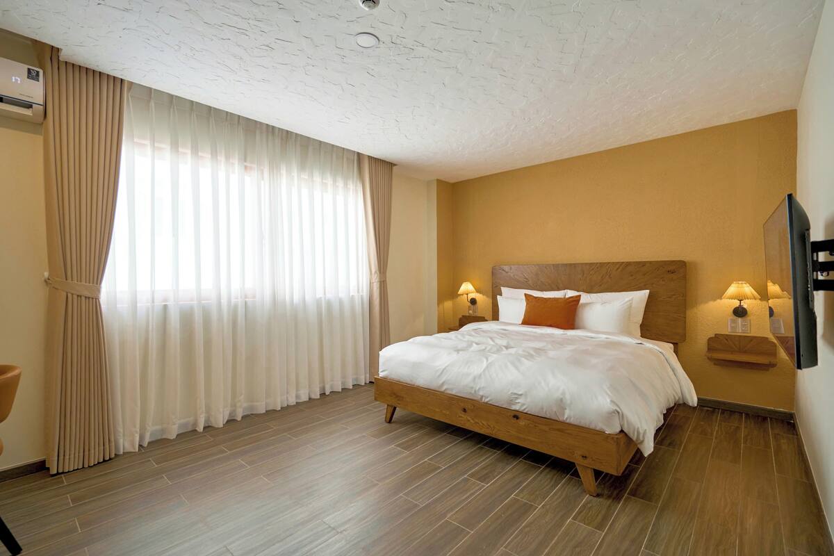 A bedroom with a wooden bed, beige curtains, a wall-mounted TV, and a ceiling fan.