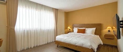 Ein Schlafzimmer mit einem hölzernen Bett, beigen Vorhängen, einem an der Wand montierten Fernseher und einem Deckenventilator.