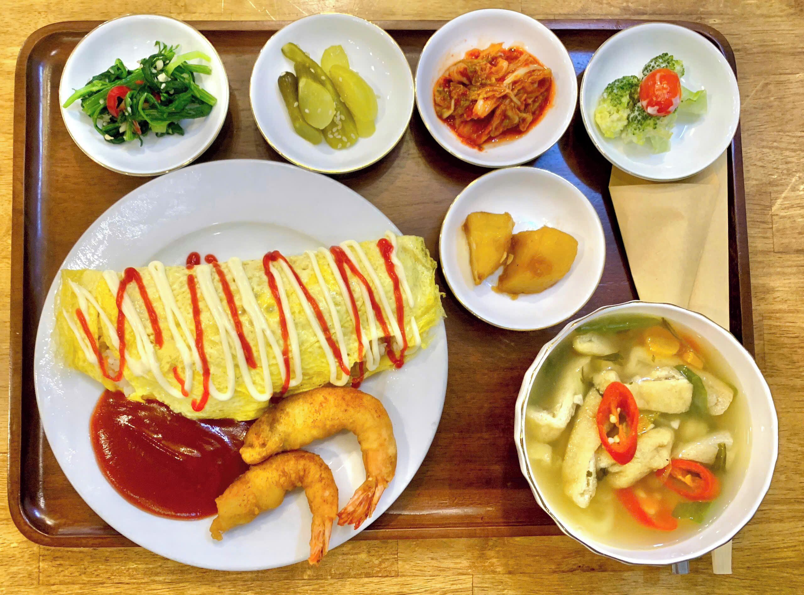 Una bandeja con diversos platos que incluyen una tortilla, camarones, kimchi, pepinillos y una sopa.