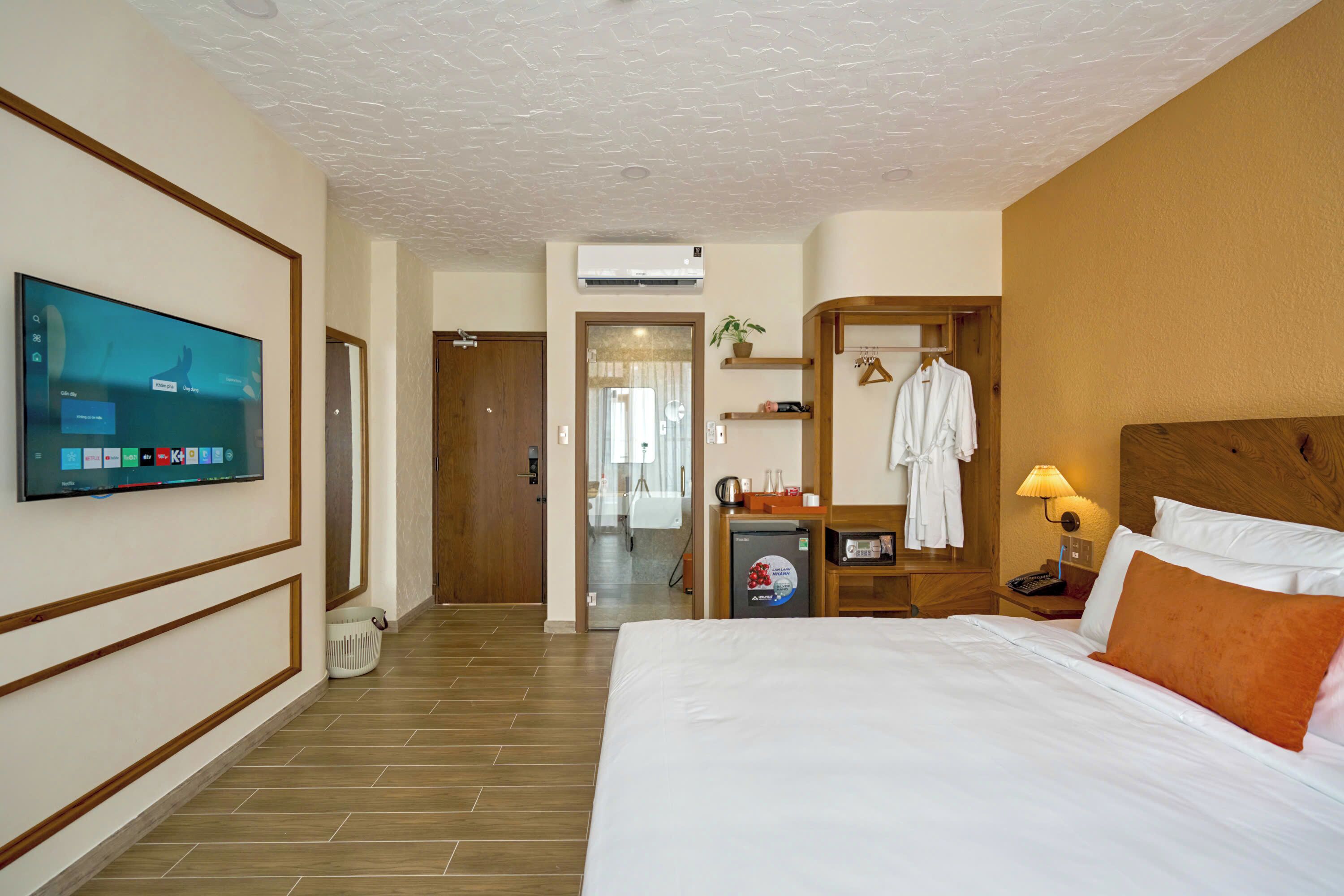 Habitación de hotel con una cama grande, televisión montada en la pared, minibar, escritorio con lámpara y cabecero de madera.