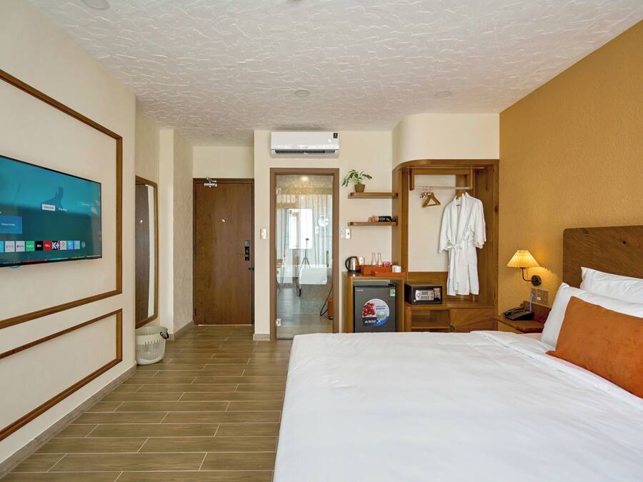 Habitación de hotel con una cama grande, televisión montada en la pared, minibar, escritorio con lámpara y cabecero de madera.