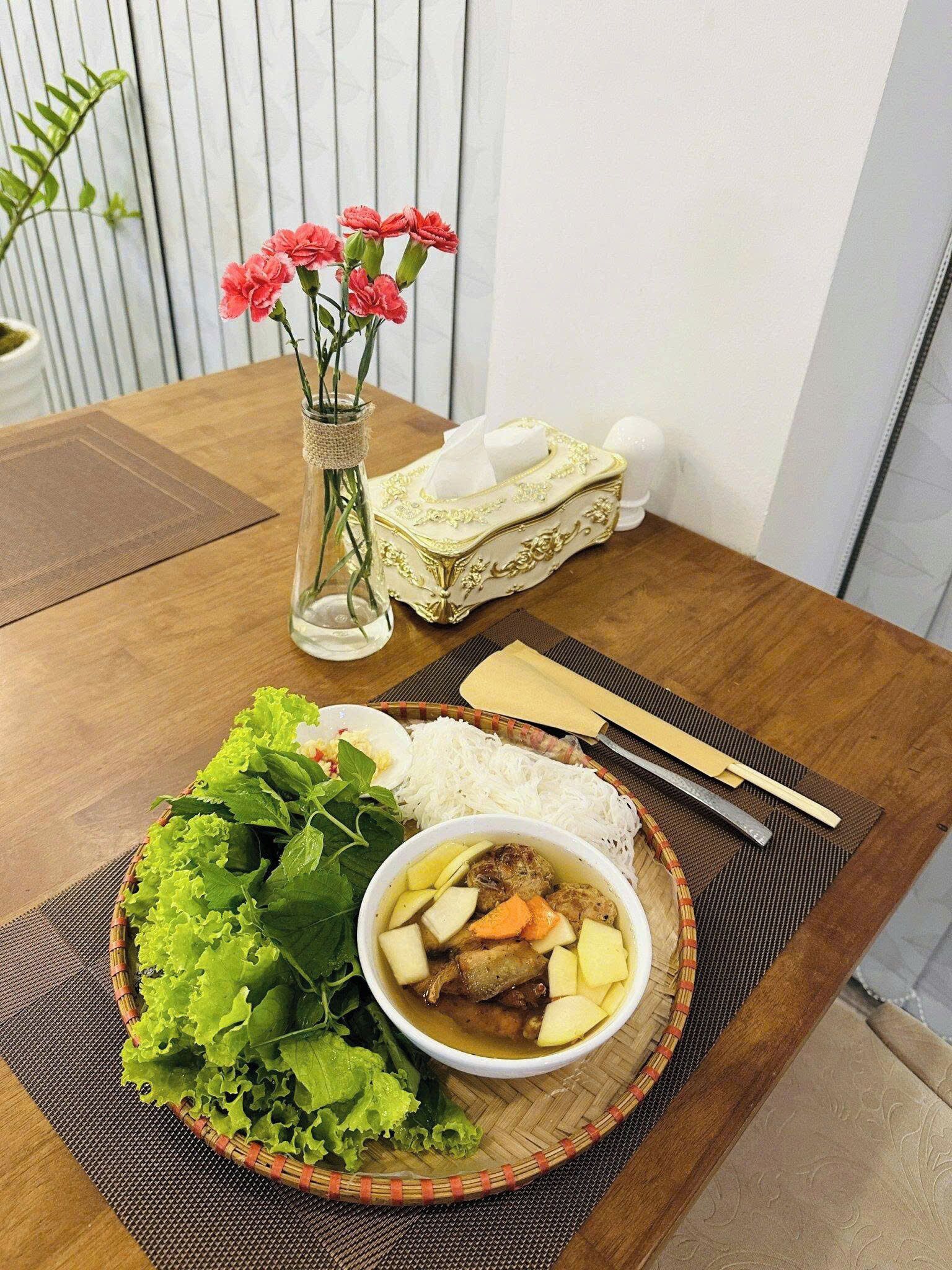 A dining table with a meal consisting of rice, a bowl of soup with meat and vegetables, a plate of fresh greens, and a vase with pink flowers.