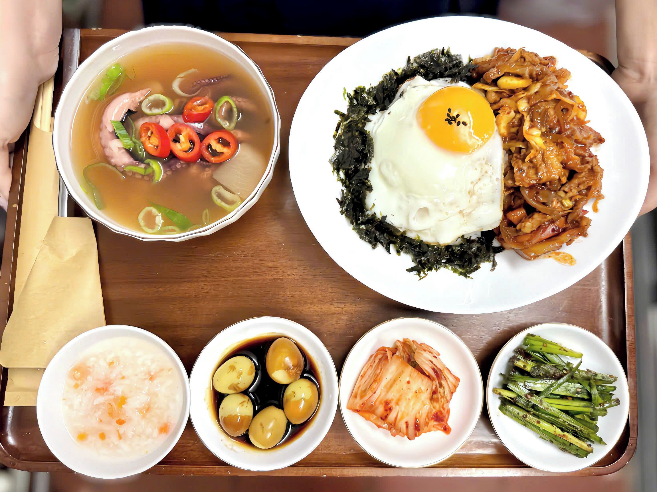 A tray with a bowl of soup, a plate with a fried egg and meat, and several small dishes of side items.