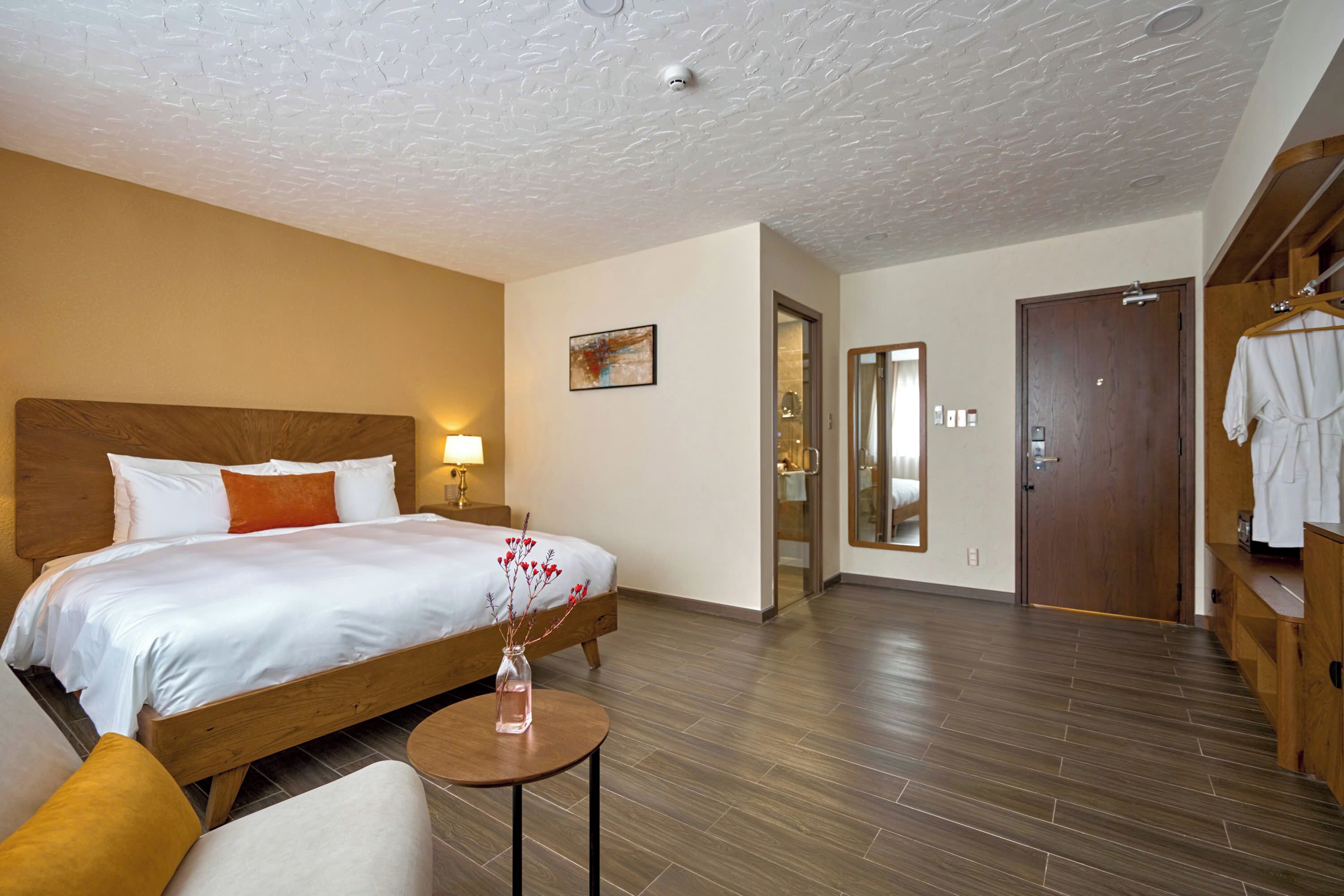 Une chambre d’hôtel avec un lit en bois, une petite table sur laquelle est posé un vase, une armoire et une porte donnant sur une salle de bain.
