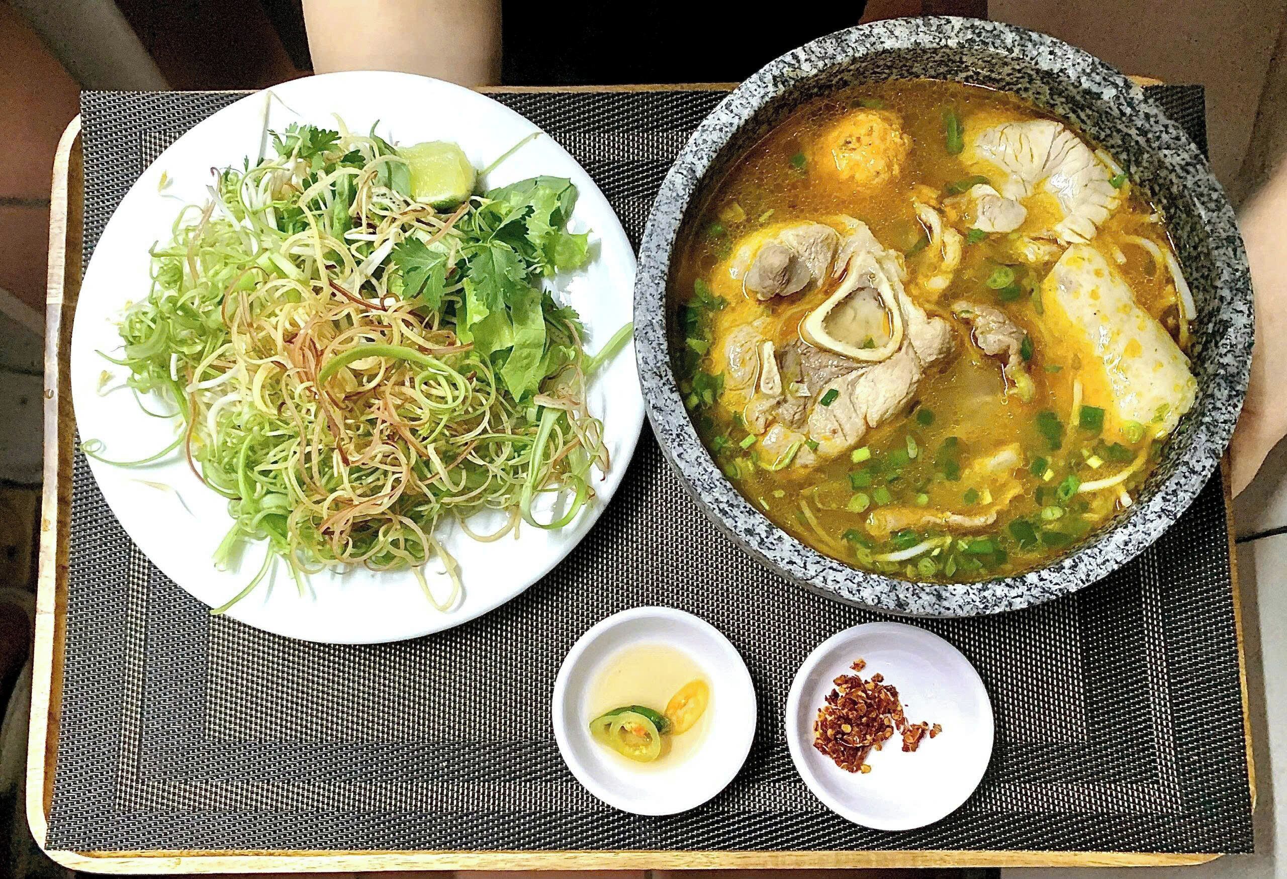 A meal consisting of a bowl of soup with meat and vegetables, a plate of noodles with greens and lime, and two small bowls of condiments.