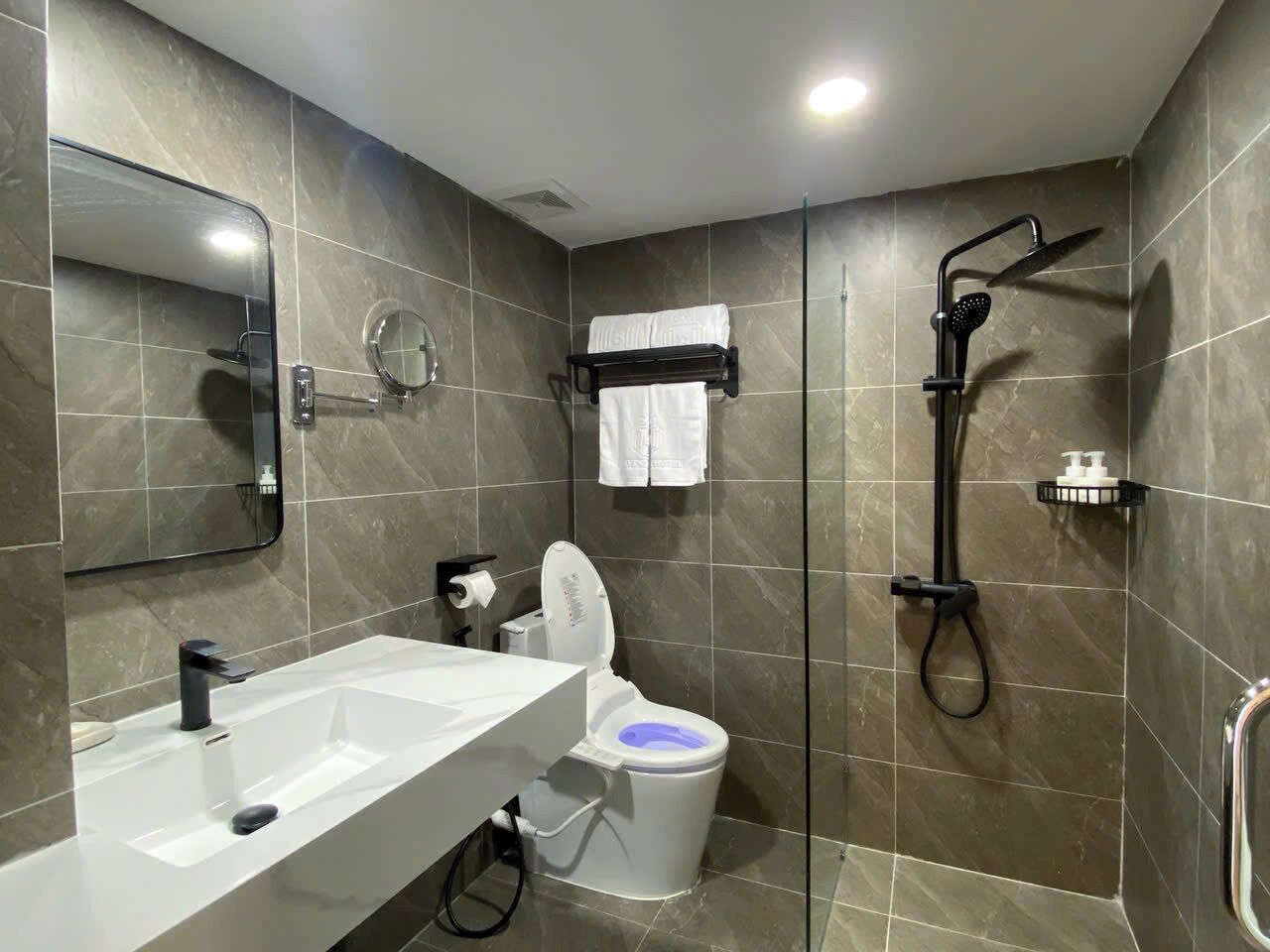 Ein modernes Badezimmer mit einem weißen Waschbecken, einer Toilette mit blauem Sitz, einer Dusche mit Duschbrause und einem Handtuchhalter mit einem weißen Handtuch.