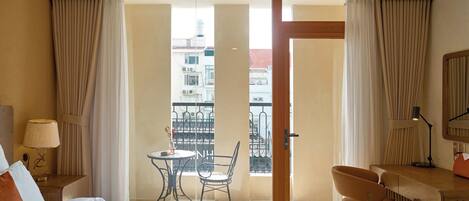 A hotel room with a bed, a desk, a chair, a table, and a balcony with a view of buildings.