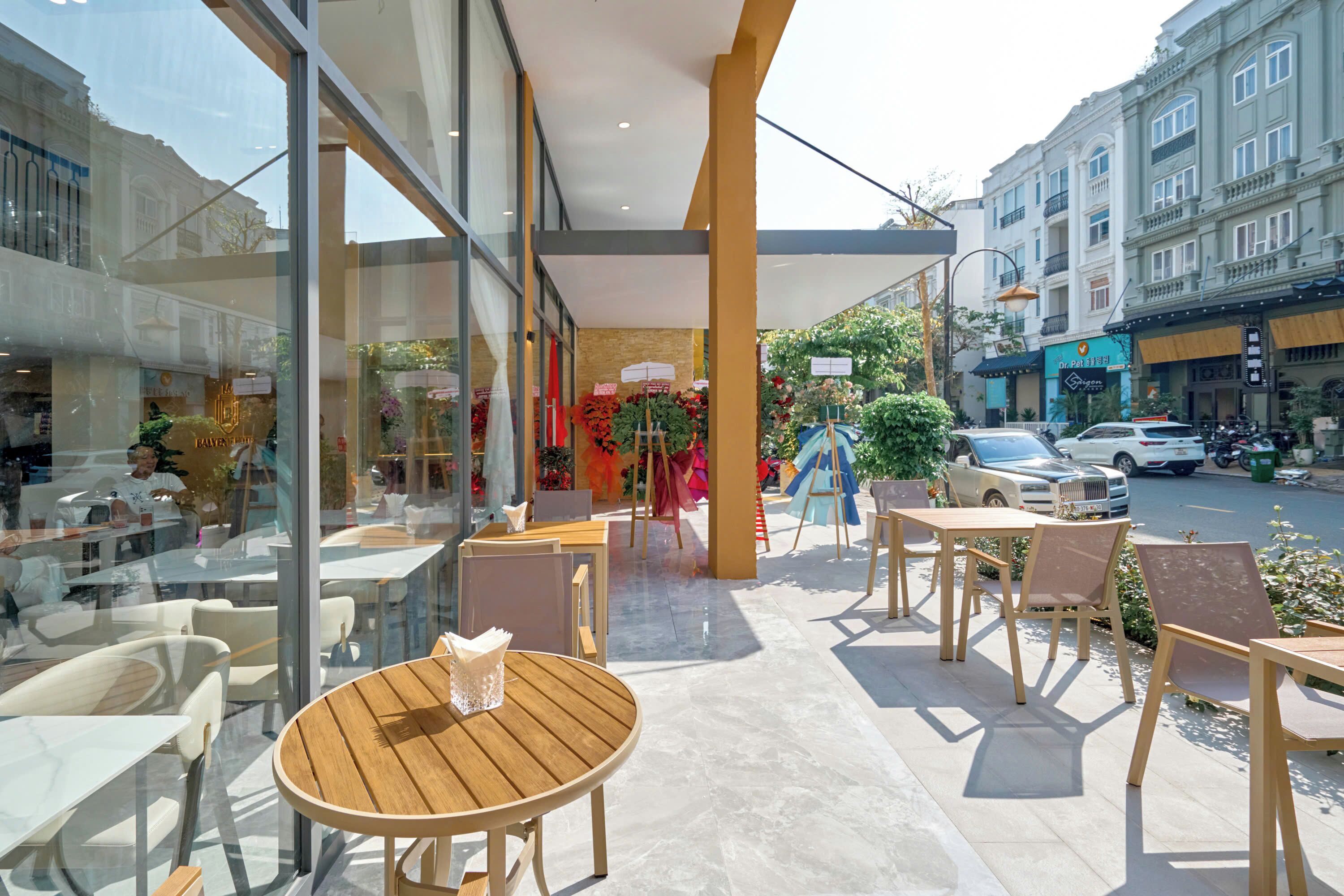 An outdoor cafe with round wooden tables and chairs, large windows, and a view of the street.