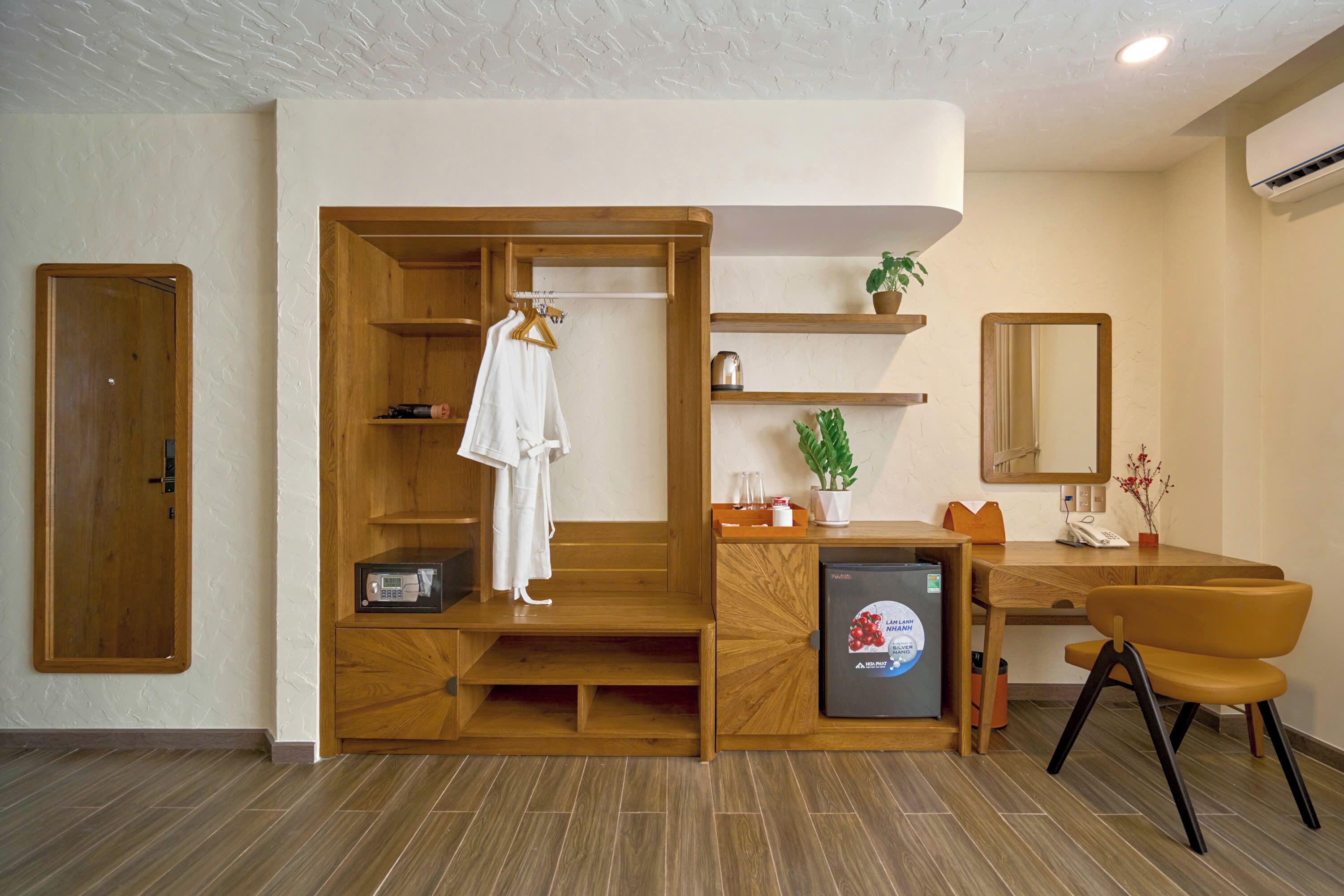 A compact hotel room with a wooden wardrobe, a small desk with a chair, a refrigerator, a mirror, and a potted plant.