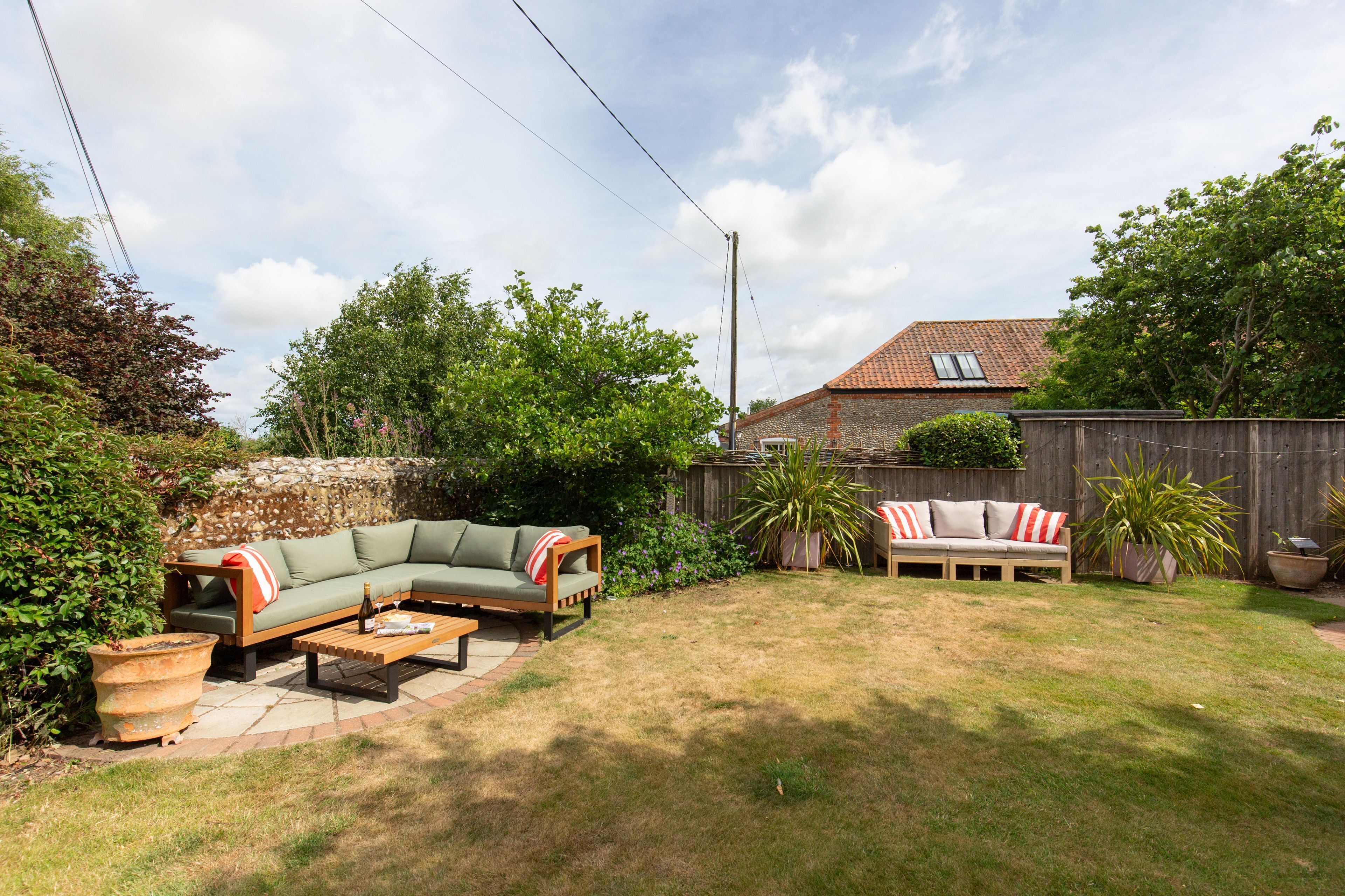 A backyard with a green sectional sofa, a wooden coffee table, and a brick patio. There are potted plants and a wooden fence in the background.