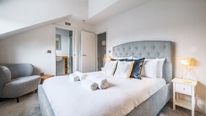 Een keurig opgemaakt bed met opgerolde handdoeken, een grijze gestoffeerde fauteuil, een nachtkastje met een lamp en een deur naar een badkamer.