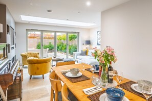 A dining area with a wooden table set for two, a piano, and a sofa in the background. - The Albans, Burnham Market, Norfolk (Burnham Market)