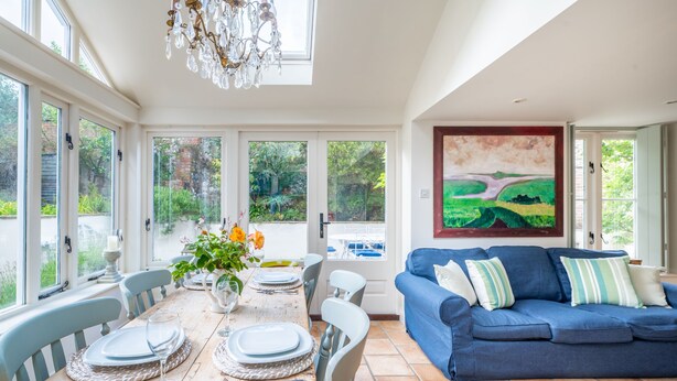 A dining area with a table set for four, a blue sofa, and a large landscape painting on the wall. - Littlefold, Brancaster, Norfolk (Brancaster)