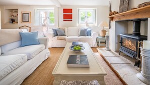 A cozy living room with a white sofa, a coffee table with a book and a bowl of fruit, a fireplace, and a window with a view of greenery. - Littlefold, Brancaster, Norfolk (Brancaster)