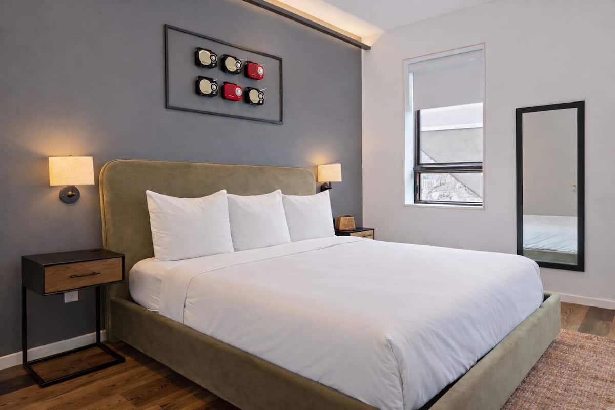 A modern hotel room with a large bed, two bedside tables, a wall-mounted artwork, a window, and a mirror.