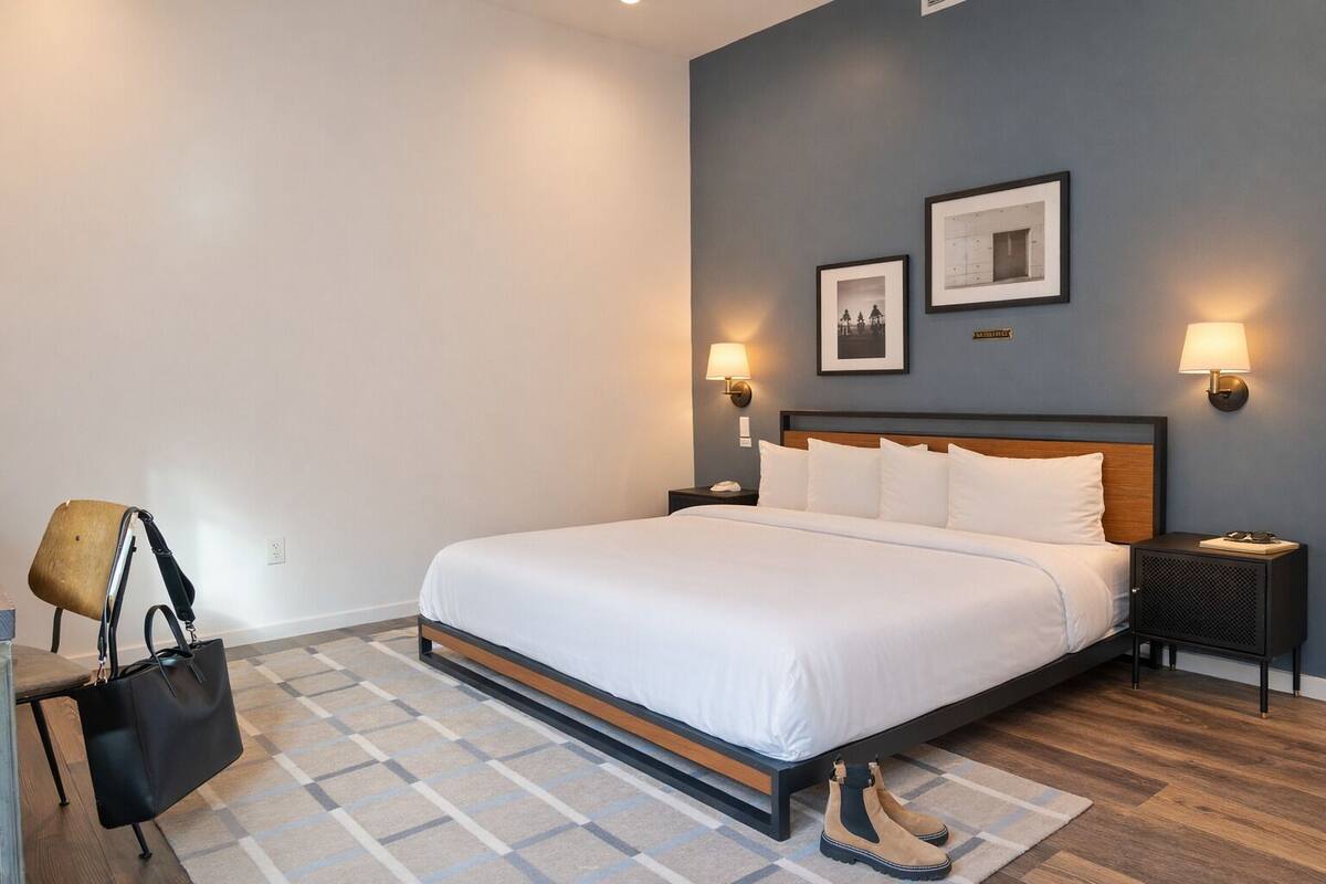 A hotel room with a bed, nightstands, wall-mounted lamps, a chair, a handbag, and a pair of boots.