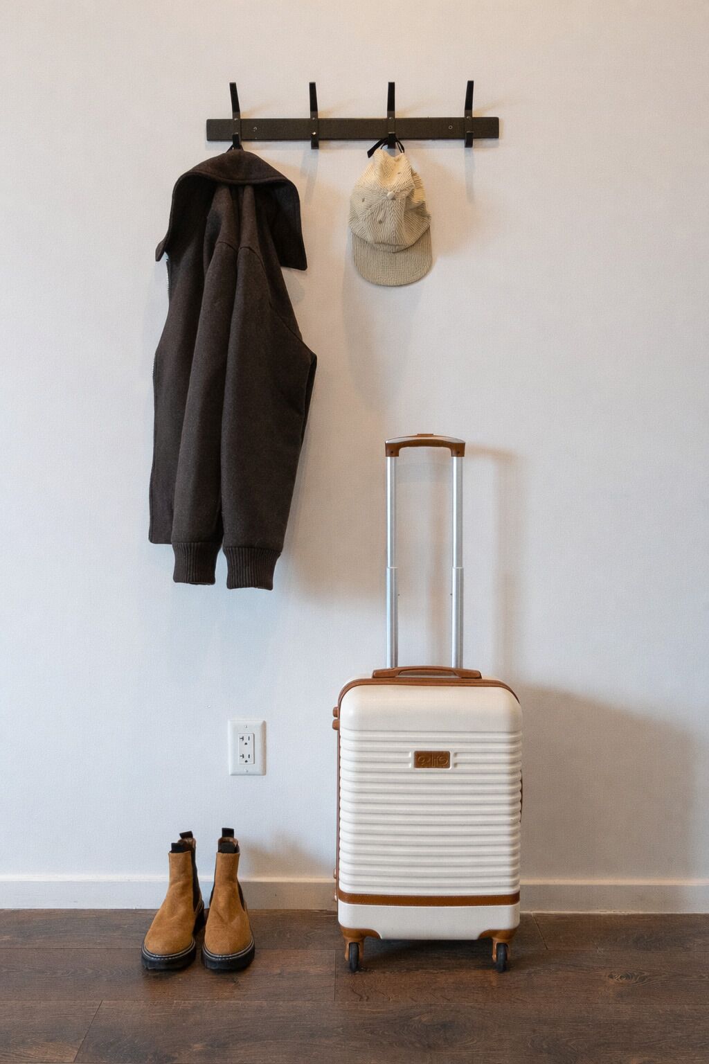 Ein Zimmer mit an der Wand montiertem Kleiderhaken, an dem eine Jacke und ein Hut hängen. Ein rollbarer Koffer und ein Paar Stiefel stehen auf dem Boden.