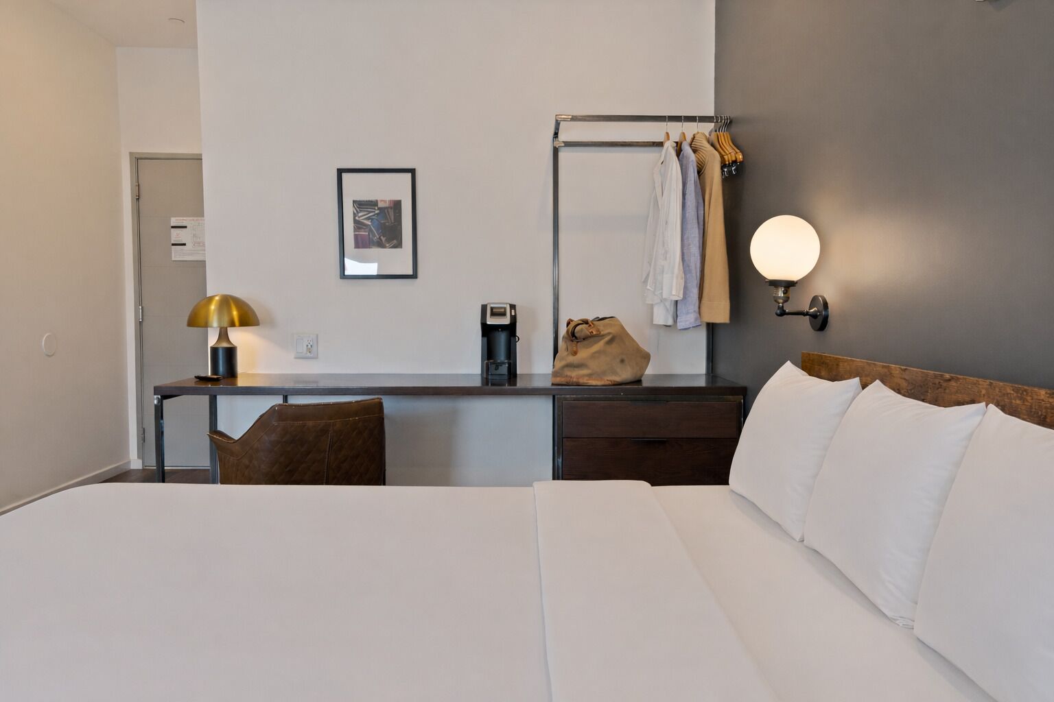 A hotel room with a bed, a desk with a lamp, a coffee maker, and a coat rack with clothes.