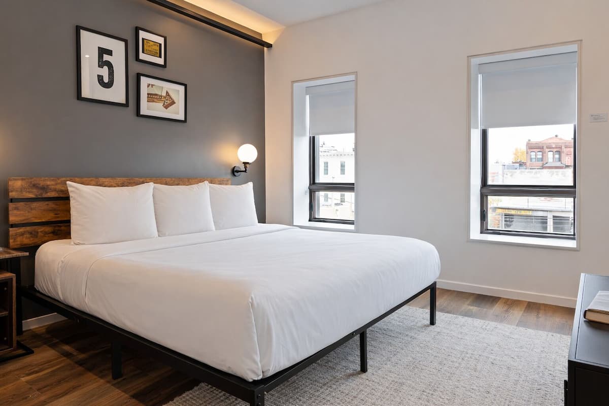A bedroom with a bed, a nightstand, a wall-mounted light, a window, and framed pictures on the wall.