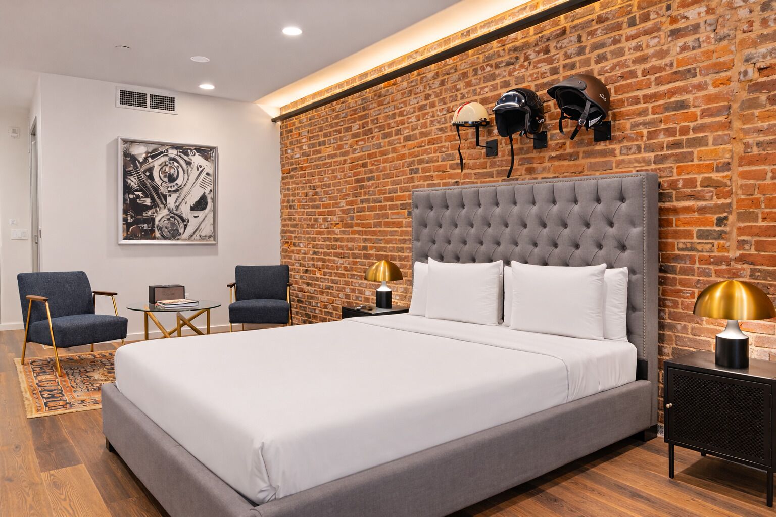 A modern bedroom with a grey tufted headboard, a large bed, two armchairs, a small side table, and a brick wall.