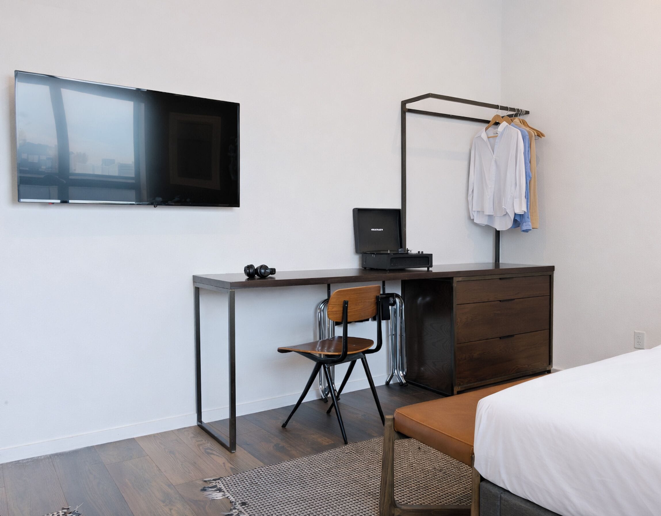 A bedroom with a bed, a desk with a chair, a TV mounted on the wall, and a clothing rack.