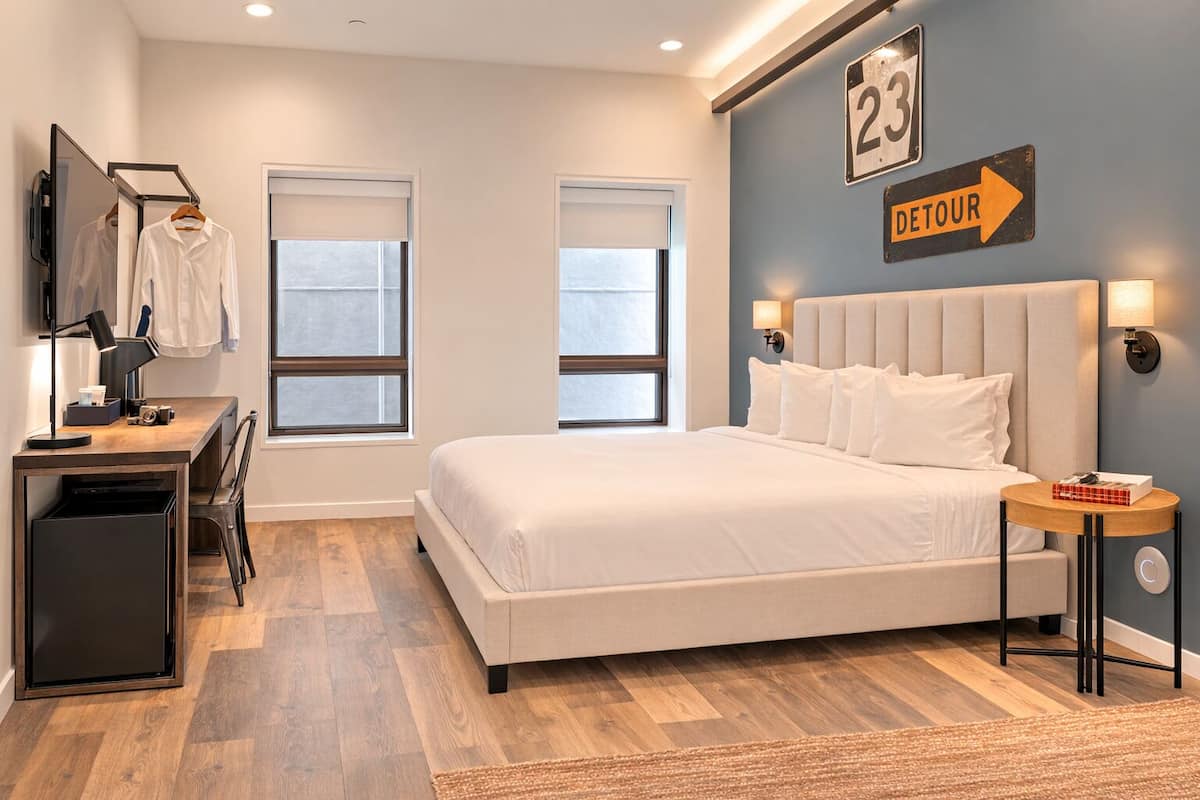 A bedroom with a bed, bedside tables, a desk, and a chair. There are two windows, a wall clock, and a sign with the number 23 and the word 'DETOUR'.