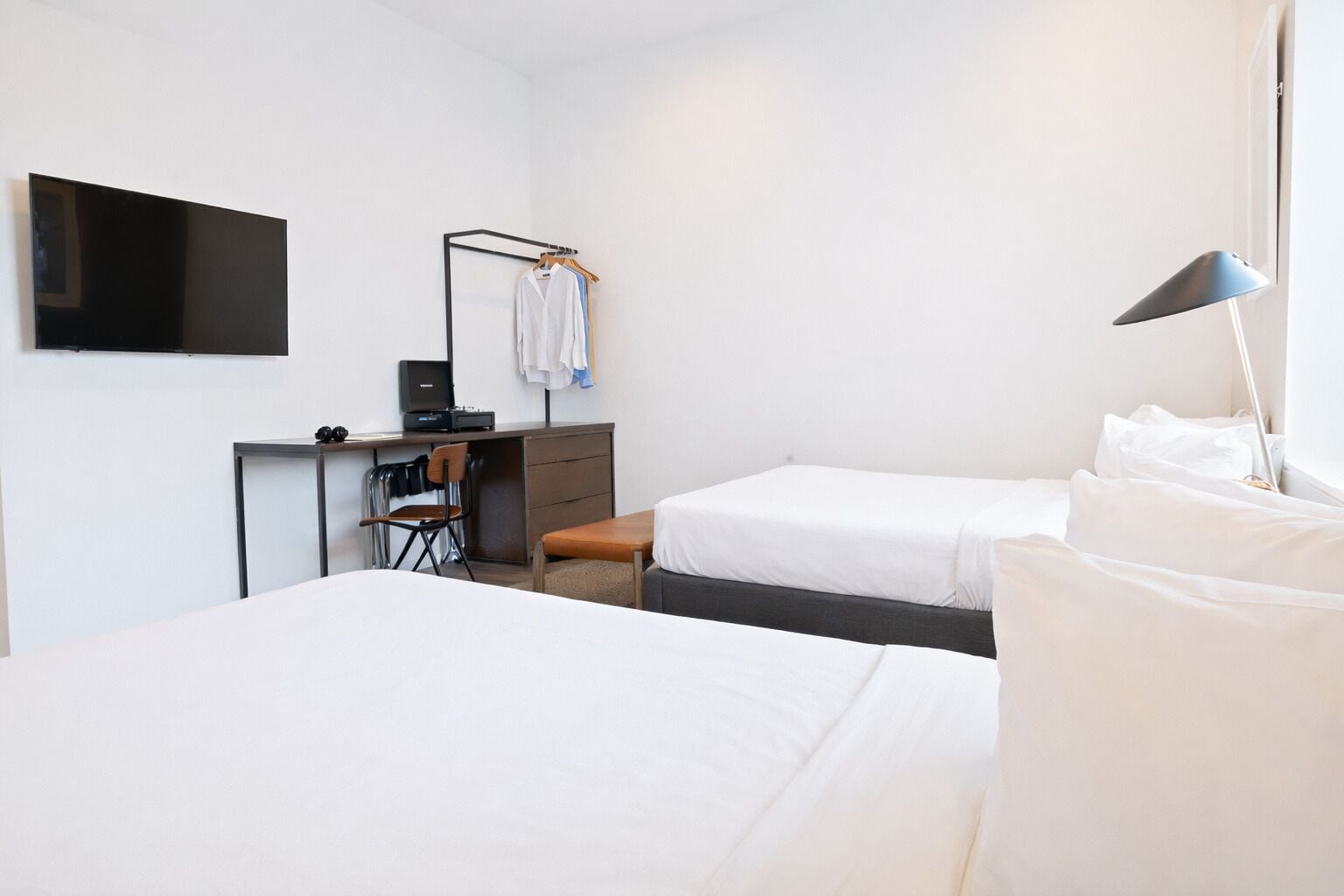 Habitación de hotel con dos camas, un escritorio con silla, un televisor montado en la pared y una lámpara.