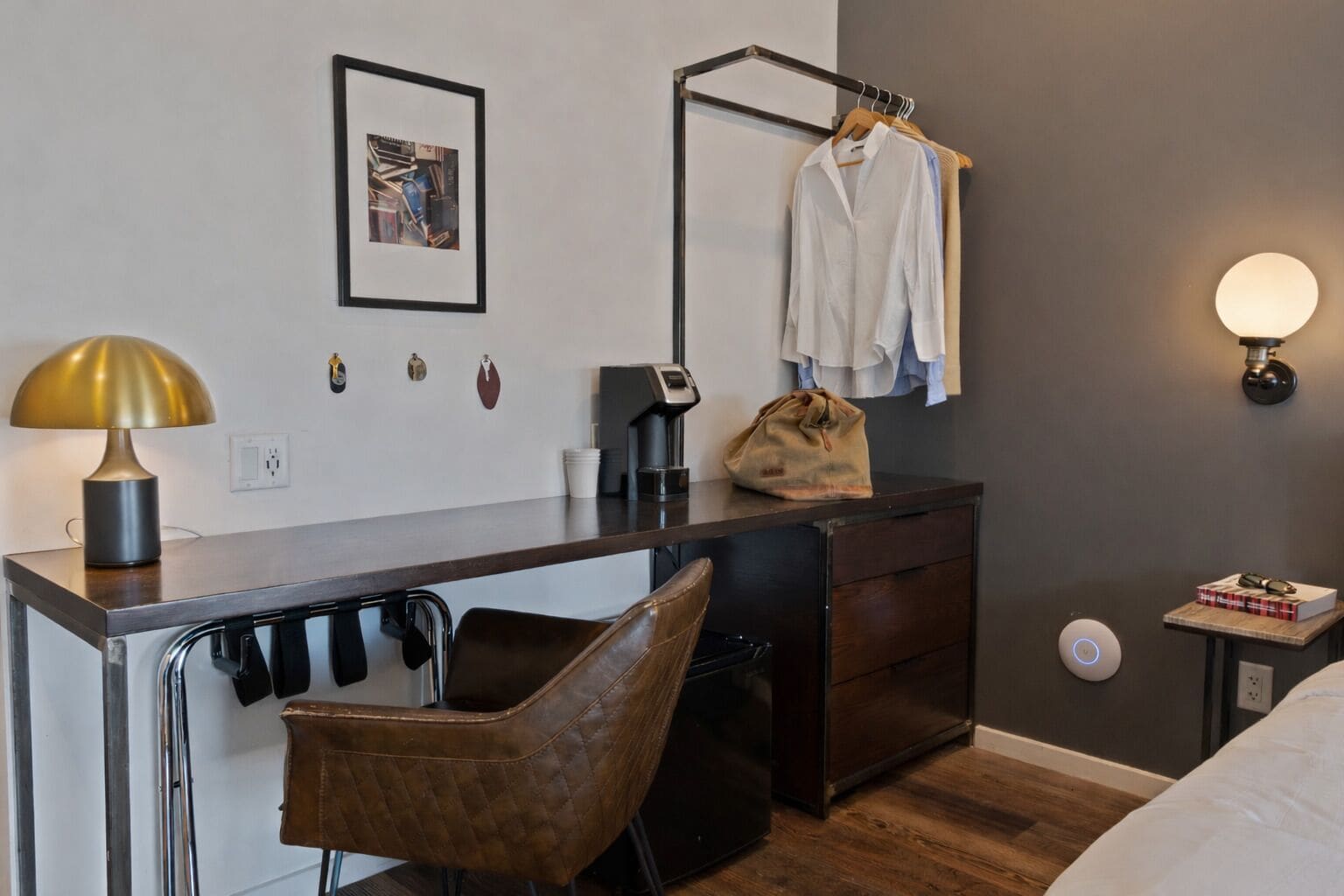 Habitación de hotel moderna con escritorio, silla, mesita de noche, lámpara y toallero en la pared.