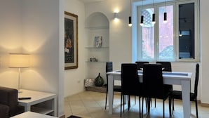 Una sala de estar moderna con comedor, una mesa blanca, sillas negras, una lámpara, un cuadro enmarcado y una ventana con vista a árboles.