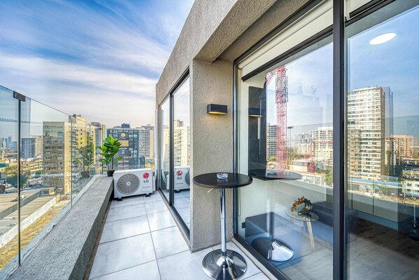A modern balcony with a glass railing, a small table, a potted plant, and an air conditioning unit.
