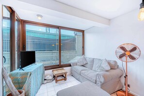 Ein Wohnzimmer mit großem Fenster, Flachbildfernseher, einer Couch, einem Standventilator und einem kleinen Holztisch.