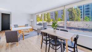 Una habitación moderna con cama, mesa de comedor, sillas, una planta en maceta y ventanas grandes.