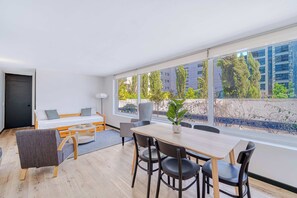 Una habitación moderna con cama, mesa de comedor, sillas, una planta en maceta y ventanas grandes.