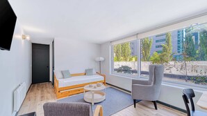 Una habitación moderna con cama, una silla, una mesa y un ventanal con vistas a edificios y árboles.
